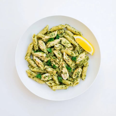 Grilled chicken pieces and penne pasta tossed in a vibrant green chimichurri sauce in a white bowl.